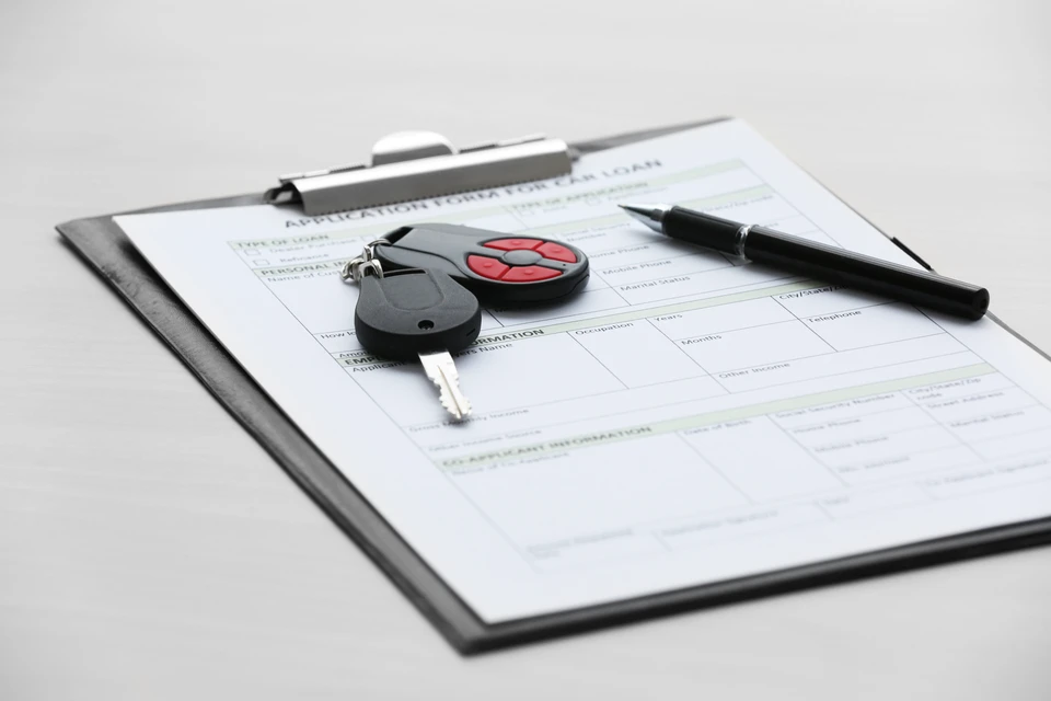 Porte-documents avec formulaire de demande de crédit automobile partiellement rempli. Clés de voiture avec télécommande rouge et stylo noir posés sur le document. Surface blanche neutre.