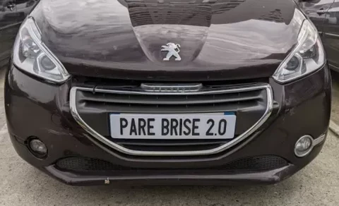 Scène extérieure avec véhicule Peugeot noir stationné sur pavés. Plaque d'immatriculation personnalisée "PARE BRISE 2.0" visible à l'avant.