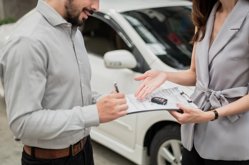 Homme en chemise claire signant un contrat automobile avec une femme en tailleur beige dans un showroom. Elle présente des documents incluant une clé de voiture. Véhicule blanc visible en arrière-plan.