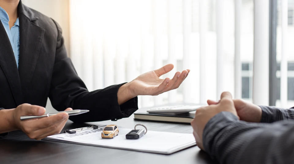 Réunion d'affaires dans bureau moderne avec deux personnes discutant autour d'une table. Une personne en costume sombre gesticule explicativement face à un client. Documents contractuels, petite voiture miniature jaune et clés automobile disposés sur la surface de travail suggèrent une transaction de vente véhicule.