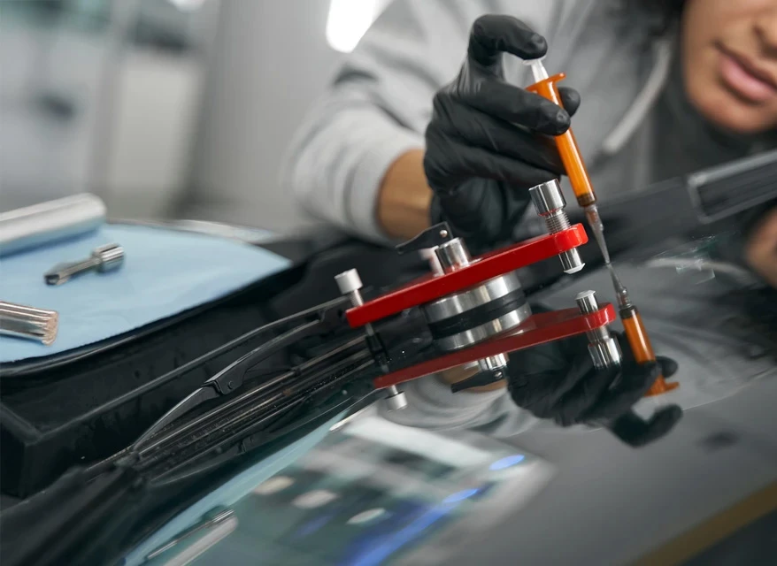 Deux techniciens remplacent un pare-brise de voiture dans un atelier vitrage automobile. L'homme de gauche porte une casquette noire, celui de droite un t-shirt bleu. Ils manipulent des ventouses de pose sur une berline noire.