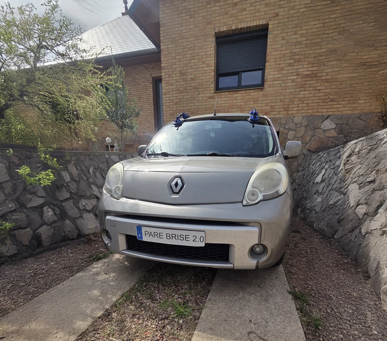 Véhicule utilitaire blanc avec bandes réflectives rouges et blanches garé en extérieur. Plaque d'immatriculation "PARE BRISE 2.0" visible à l'avant.