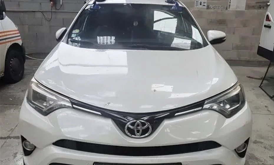 Pare-brise de voiture avec impact circulaire central et fissures radiales étendues. Véhicule blanc stationné en extérieur, reflets d'arbres visibles sur la vitre endommagée.