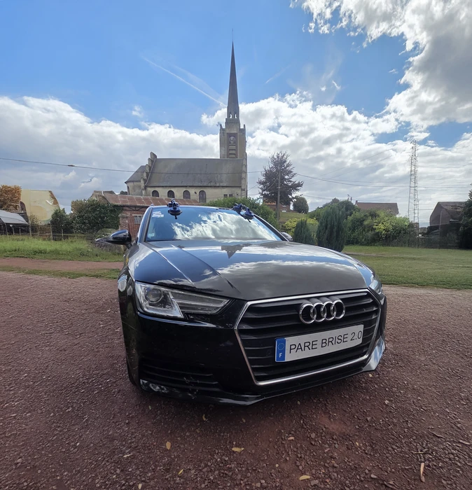 Scène extérieure résidentielle avec véhicule Renault beige stationné devant maison en briques. Plaque d'immatriculation personnalisée "PARE BRISE 2.0" visible. Environnement pavillon avec mur en pierre et végétation.