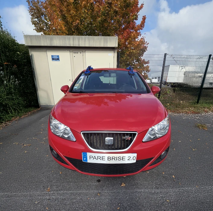 Scène extérieure résidentielle avec véhicule Renault beige stationné devant maison en briques. Plaque d'immatriculation personnalisée "PARE BRISE 2.0" visible. Environnement pavillon avec mur en pierre et végétation.