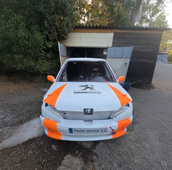 Intérieur véhicule sous la pluie, main d'homme près du volant face à un pare-brise fissuré. Impact central en étoile avec lignes de fracture radiales sur vitre avant mouillée, gouttes d'eau visibles.
