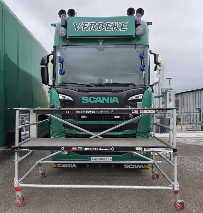 Atelier de réparation automobile, mécanicien en combinaison rose travaillant sur roue de camion. Intervention sur pneumatique poids lourd monté sur jante alliage, utilisation d'équipement de démontage hydraulique rouge. Environnement professionnel avec sols métalliques antidérapants, outillage spécialisé transport routier.