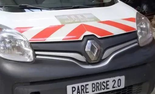 Véhicule utilitaire blanc avec bandes réflectives rouges et blanches garé en extérieur. Plaque d'immatriculation "PARE BRISE 2.0" visible à l'avant.